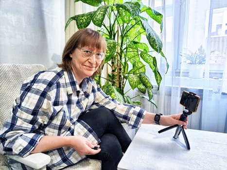 Smiling woman in glasses using a smartphone on a tripod for a video chat. A middle-aged woman posing and taking a selfie
