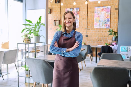 Confident successful young woman service worker owner in apron with crossed arms looking at camera in restaurant cafeteria coffee pastry shop interior. Small business staff occupation entrepreneur work