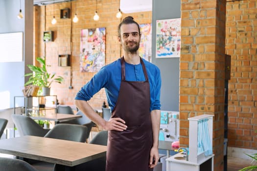 Confident successful young man service worker owner in apron, handsome male looking at camera in restaurant cafeteria coffee pastry shop interior. Small business staff occupation entrepreneur work