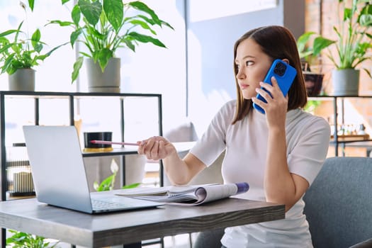 Young woman sitting at table with laptop computer talking on mobile phone in coworking cafe. Female university college student preparing for test exam, freelancer, business employee working remotely