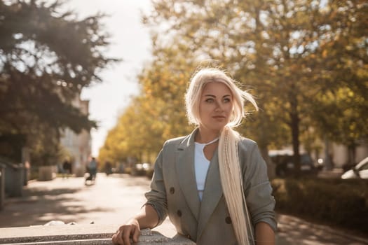 A blonde woman wearing a gray jacket and white shirt stands on a sidewalk. She is looking at the camera with a smile on her face