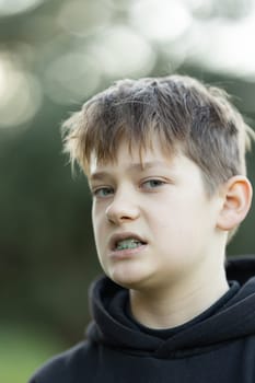 A boy with a green mouth is standing in a park. He is wearing a black hoodie and has a green mouth guard