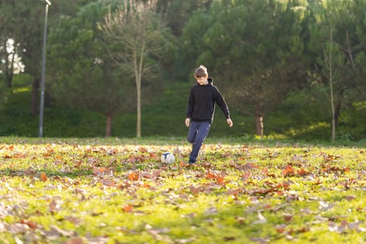 A boy is kicking a soccer ball in a field. The boy is wearing a black hoodie and blue pants. The field is covered in leaves, giving it a fall-like atmosphere