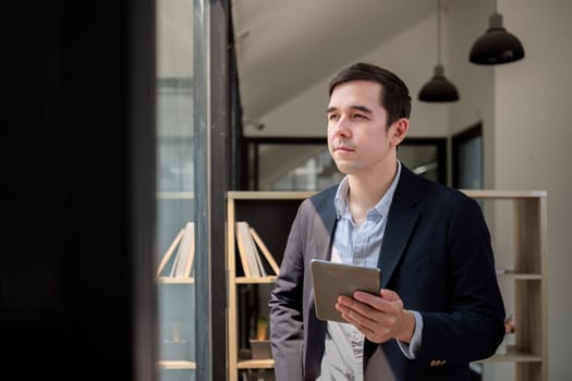 Happy young businessman using online app on tablet for job communication, looking out the window at office.