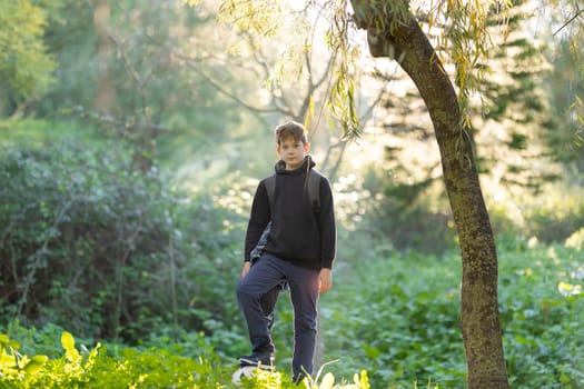 A boy is standing in a forest with a backpack on. The boy is wearing a black hoodie and blue pants