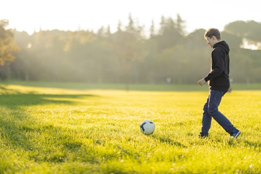 A boy is playing soccer in a field. The grass is green and the sky is blue. The boy is wearing a black hoodie and blue jeans