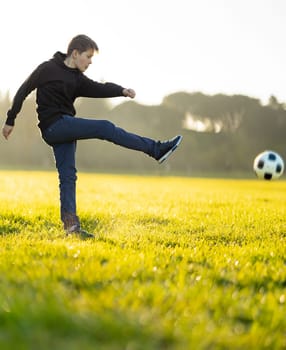 A boy kicks a soccer ball in a field. The boy is wearing a black hoodie and blue jeans