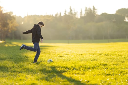 A boy kicks a soccer ball in a field. The boy is wearing a black jacket and blue jeans. The field is lush and green, and the sun is shining brightly. Scene is cheerful and active