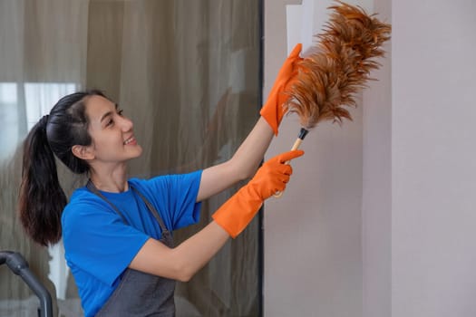 A beautiful young housekeeper does housework wearing orange rubber and uses a duster to clean up the items in the room..