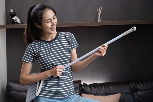 Cheerful and happy young woman singing and dancing while cleaning the living room, holding house cleaning equipment and wearing headphones, singing happily..