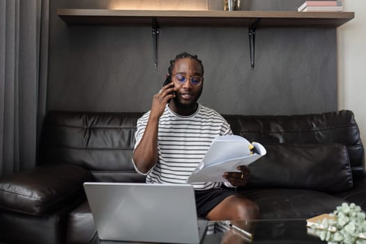 African American man working with document and laptop while talking on mobile at home on couch. work from home concept.