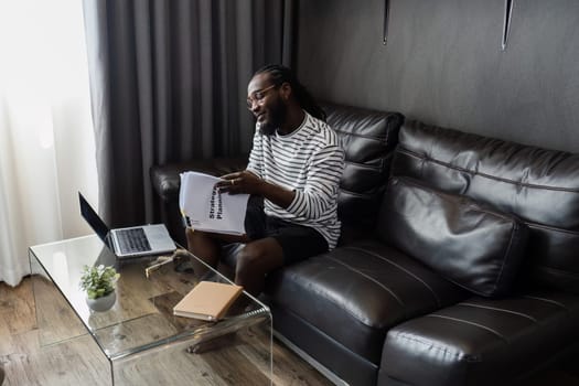 African American man working with document and laptop while talking on mobile at home on couch. work from home concept.