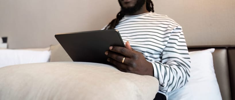 Smiling young African American man using digital tablet remote job sitting on the bed at home. concept online technology communication.