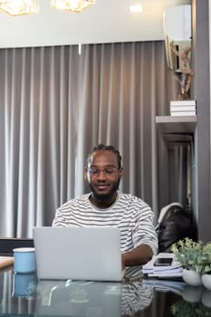 African American man working with laptop computer remote while sitting at glass table in living room. Black guy do freelance work at home office.