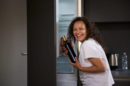 Young curly haired brunette woman with alcohol addiction, holding a bottle of wine and drinking alcohol in the morning, smiling looking at camera. Alcoholism, mental health and social problems