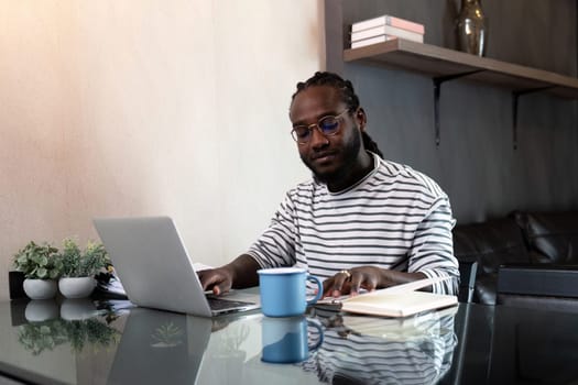 African American man working with laptop computer remote while sitting at glass table in living room. Black guy do freelance work at home office.