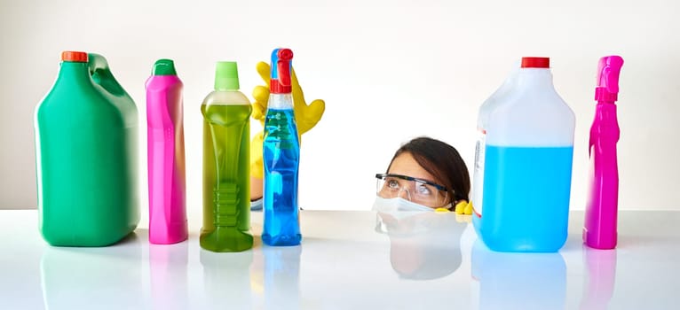 Detergent, products and woman with mask for cleaning or glasses for protection, safety and soap at home. Housekeeping, cleaner or female person with bottles, liquid container or spray on table.