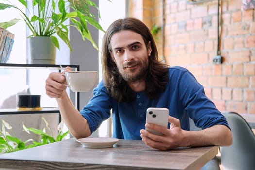 Young 30s handsome bearded with long hair man using smartphone drinking cup of coffee. Smiling guy looking at camera sitting in cafe. Mobile technologies apps for leisure work business communication