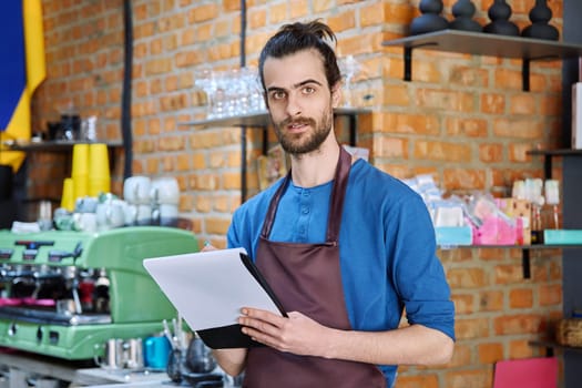 Young man in apron, food service worker, small business owner entrepreneur with work papers looking at camera near counter of coffee shop cafe cafeteria. Staff, occupation, successful business, work