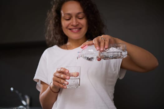 Details on a bottle of fresh clean pure water in the hands of a blurred smiling woman pouring water into drinking glass. Start your day with a sip of hydrating water for detox and diet