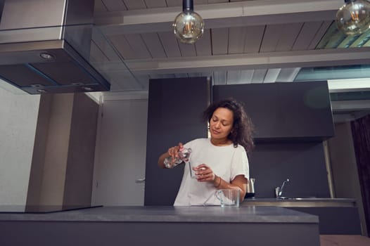 Authentic portrait of a beautiful Latin American woman standing at kitchen counter, pouring some fresh pure water into a drinking glass in the morning. Healthy lifestyle. Slimming and dieting concept