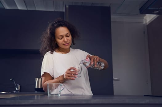 Young woman in pajamas, pouring clean fresh mineral water from bottle into a glass, drinking water in the morning while waking up for healthy diet or detox, standing at kitchen counter at home kitchen