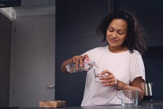 Multi ethnic smiling young woman pouring clean mineral water into a drinking glass in the morning, starting new day with healthy life habit. Healthy lifestyle and body care concept