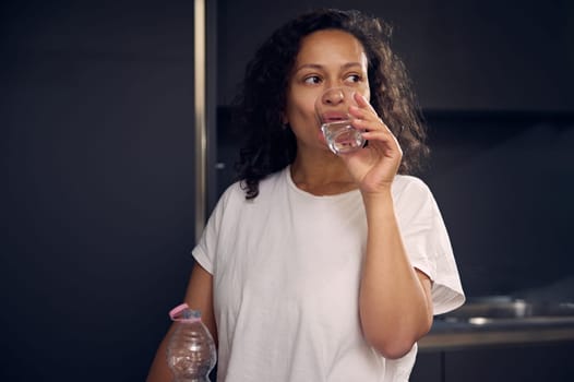 Young curly haired brunette woman in white t-shirt, drinking water in the morning, renewing aqua balance, taking care of her body, standing against modern gray color minimalist kitchen background