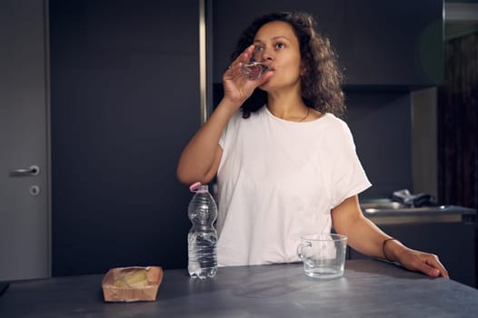 Healthy lifestyle concept. Beautiful multi ethnic curly haired young adult woman in white pajamas, standing at kitchen table and drinking pure water from glass while waking up in the morning