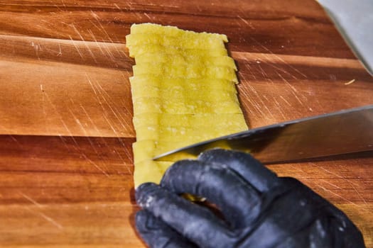 Close-up of gloved hands expertly slicing folded pasta sheets on rustic cutting board, evoking the art of food preparation in a Fort Wayne kitchen