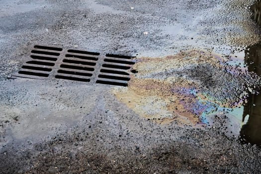 An oil or gasoline slick against the backdrop of an asphalt road flows into a storm drain. Environmental problems of water pollution.
