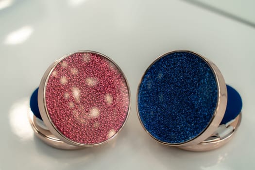 Two small round makeup compacts with glittery blue and pink designs. The compacts are placed next to each other on a white surface