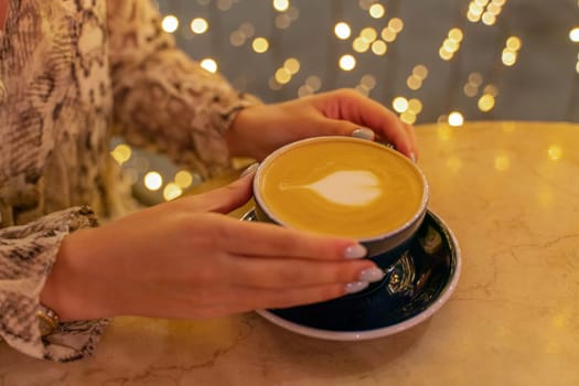 Close up cup of latte coffee in a coffee shop. Female hands hold a cup of coffee with foam latte in the shape of a heart on a table in a cafe. On the background, the lights are in blur