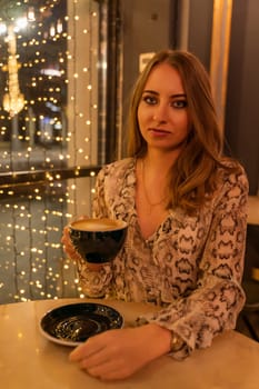 A woman is sitting at a table with a cup of coffee in front of her. She is wearing a snake print dress and she is enjoying her coffee