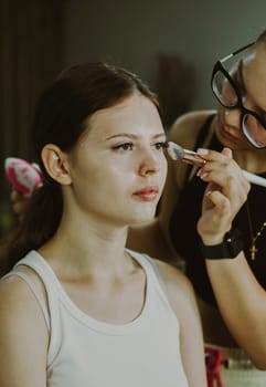 One young handsome Caucasian makeup artist applies blush with a brush to the cheekbones of a girl sitting in a chair early in the morning in a beauty salon, close-up side view. Step by step.