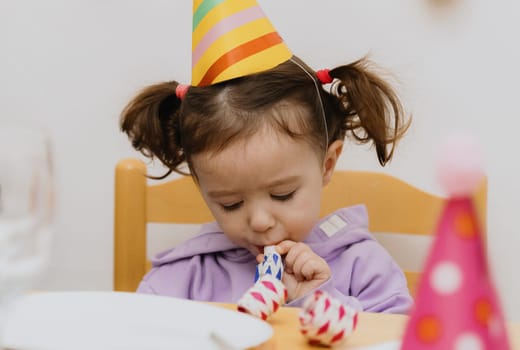 One beautiful Caucasian girl with a cone hat on her head sits at a festive table and blows into a paper tube, looking down and celebrating her birthday, close-up side view.