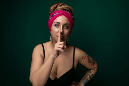 A woman wearing a pink turban is shown in the act of making a hushing gesture, with her finger pressed against her lips. She appears to be signaling for quiet or secrecy.