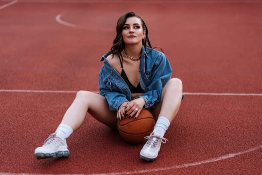 Excited brunette woman sitting at basketball court with ball between her legs. Outdoor portrait of dreaming girl in bra and jacket posing on the ground. SWAG concept.