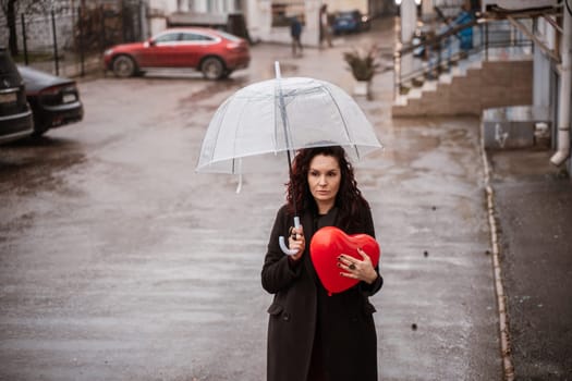A woman holding a red heart under a clear umbrella. The umbrella is open and the woman is standing in the rain