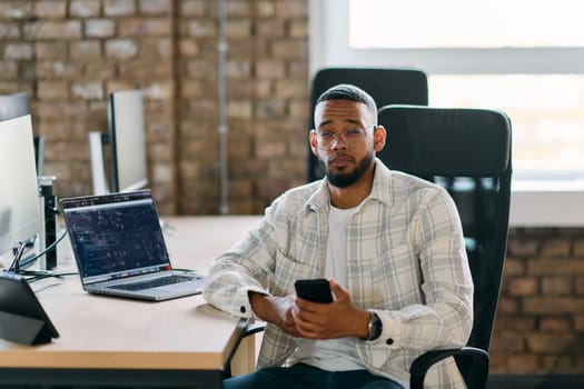 African American entrepreneur takes a break in a modern office, using a smartphone to browse social media, capturing a moment of digital connectivity and relaxation amidst his business endeavors