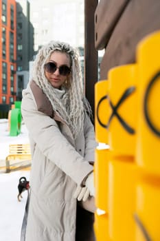 A woman with white dreadlocks stands near a yellow post in an urban setting. The woman appears relaxed and confident as she poses next to the vibrant object.
