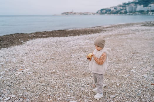 Little girl looks at a bitten apple in her hands while standing on a pebble beach. High quality photo