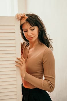 A woman is standing in front of a white door, looking at the camera. She is wearing a brown sweater and black pants. The image has a warm and cozy feeling