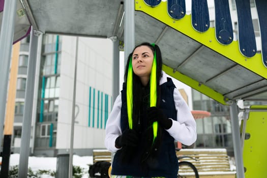 A woman is standing underneath a metal structure, with a bright yellow scarf wrapped around her neck. She appears to be observing her surroundings while standing in a urban setting.