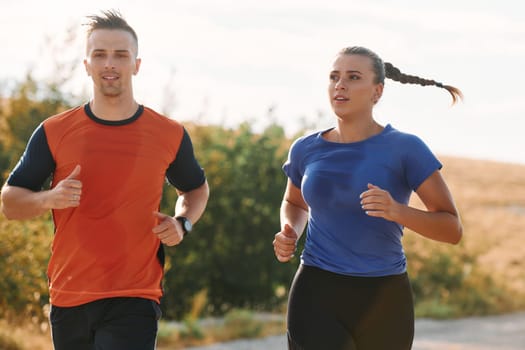 A couple dressed in sportswear runs along a scenic road during an early morning workout, enjoying the fresh air and maintaining a healthy lifestyle.