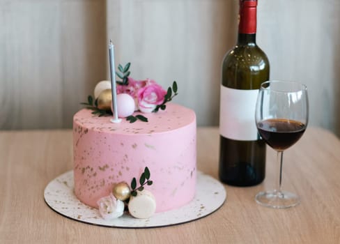 One beautiful anniversary pink cake with fresh rose flowers, a candle, a bottle of red wine and a glass are on the table, close-up side view.