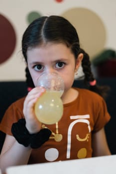 Portrait of one beautiful caucasian brunette girl drinking lemon soda from a glass while sitting at a table on a sofa in a cafe against the background of an abstract wall, close-up side view.