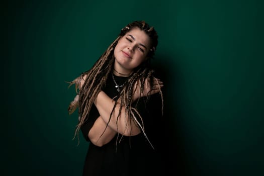 A woman with dreadlocks is standing in front of a green wall. She is looking directly at the camera, with a neutral expression. The wall provides a vibrant backdrop to the scene.