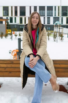 A woman is seated on a bench covered in snow, wearing a heavy coat and gloves. Snowflakes fall around her as she gazes off into the distance.