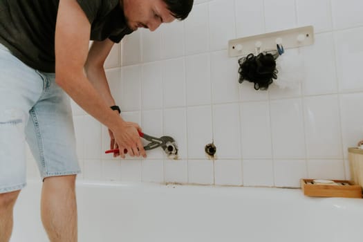 One young Caucasian recognizable guy with a brunette unscrews the pipe from the faucet with an adjustable wrench in a white tiled wall in the bathroom, close-up side view.Step by step.
